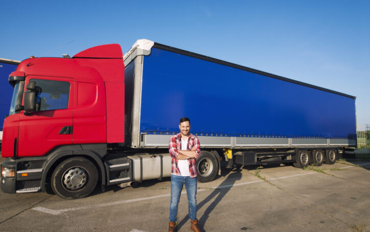 Portrait of professional American truck driver in casual clothing and boots standing in front of truck vehicle with long trailer.
