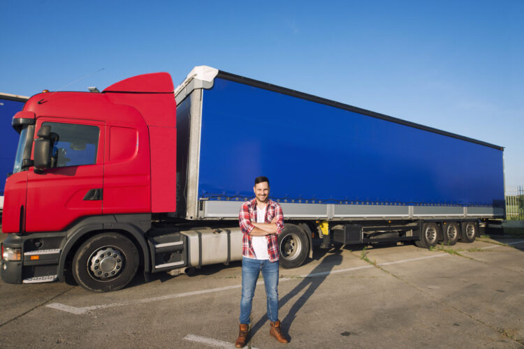 Portrait of professional American truck driver in casual clothing and boots standing in front of truck vehicle with long trailer.