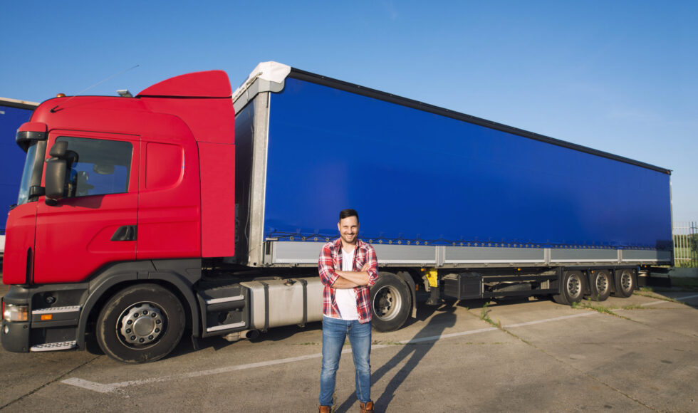 Portrait of professional American truck driver in casual clothing and boots standing in front of truck vehicle with long trailer.